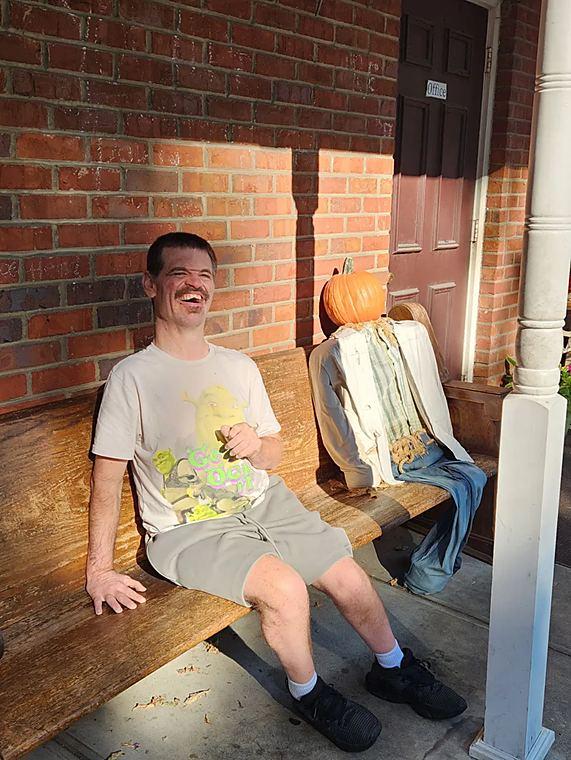 A member laughing on a bench next to a scarecrow on the porch